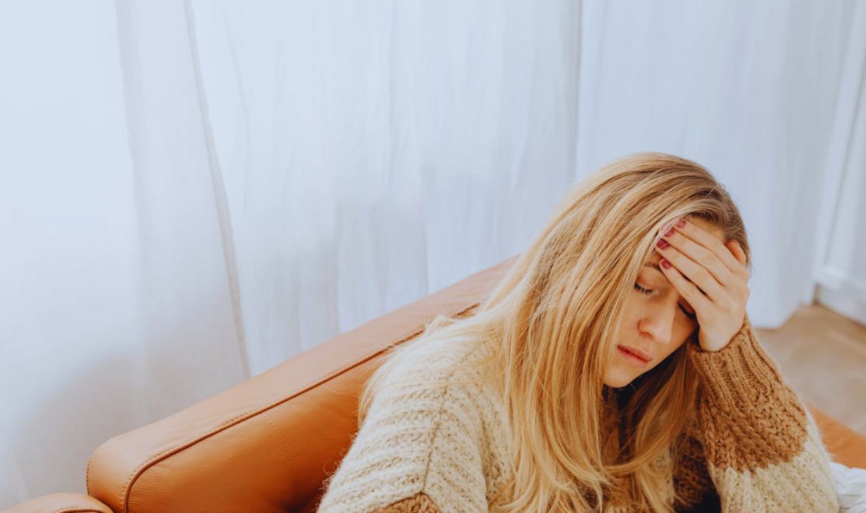 A woman in a knitted sweater sits with a headache on a cozy sofa indoors