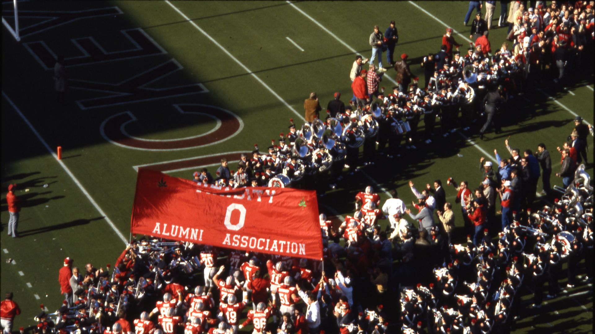 A view of the 1987 Ohio State Buckeyes football team as they run out onto the field in Ohio Stadium. Members of the Varsity O Alumni Association hold a banner for the players to run under and, along with the marching band, they form two lines for the play