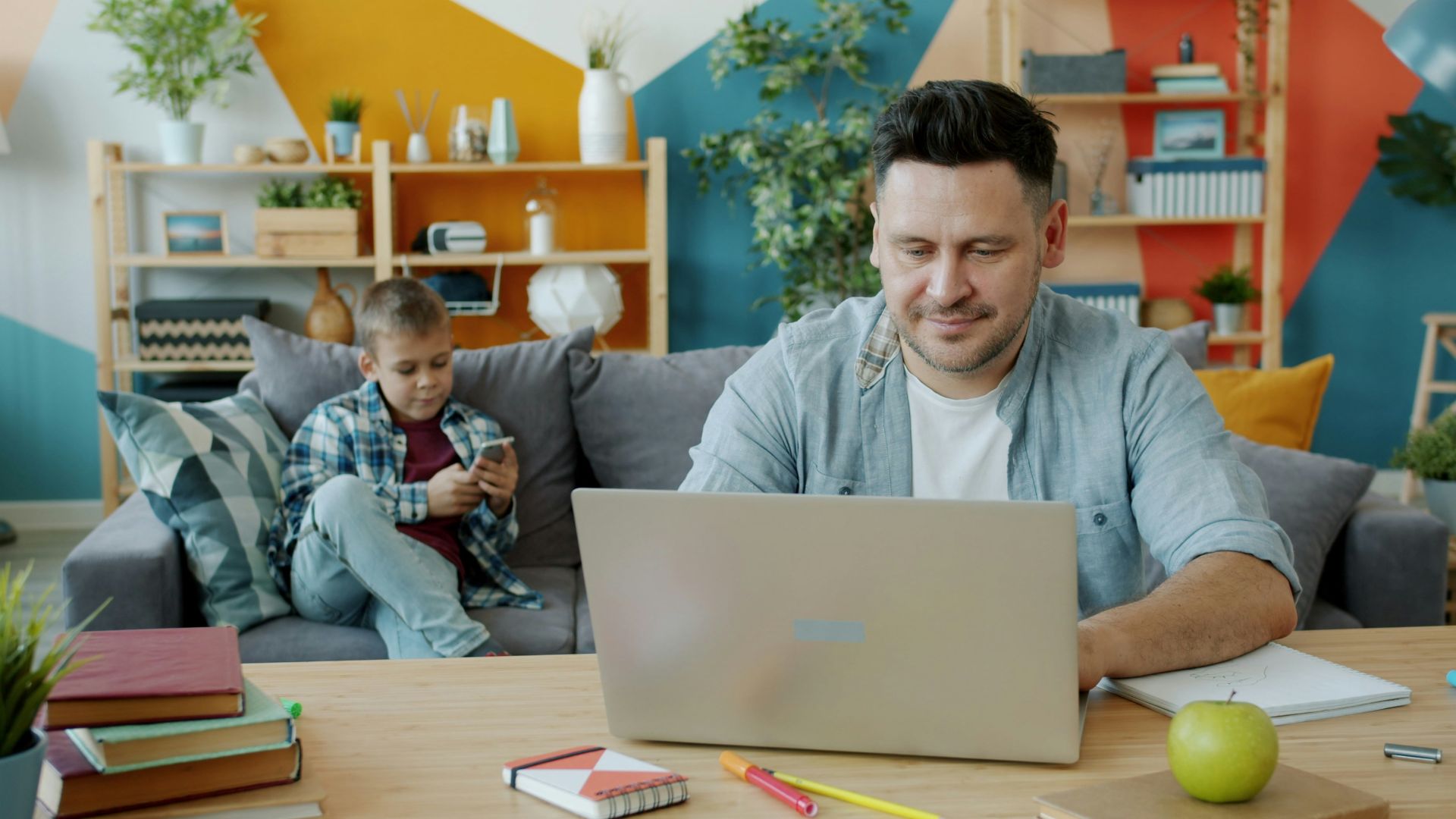 Father works on laptop while son plays on phone.