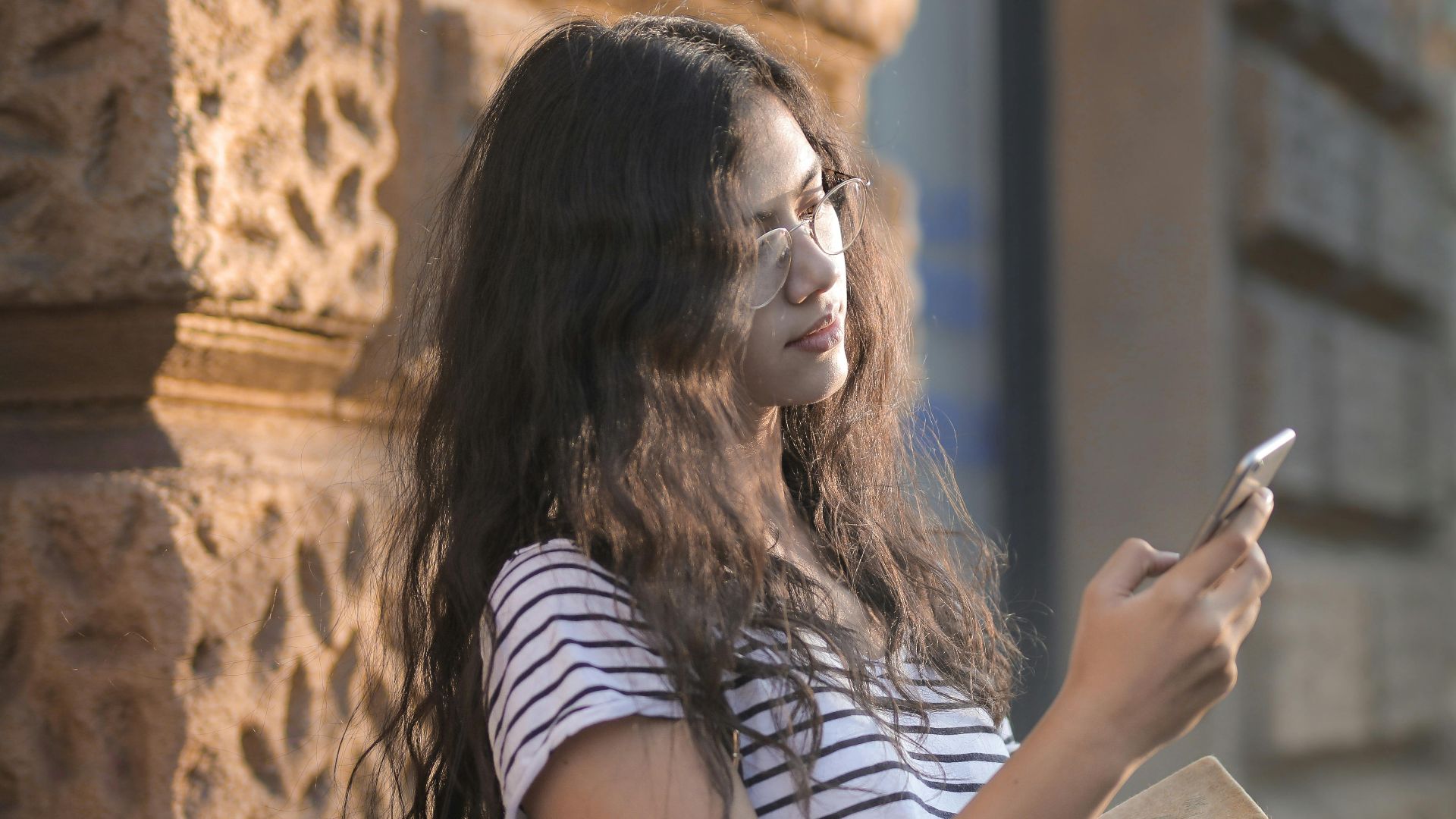 Casual portrait of a young woman leaning on a wall, using her smartphone and holding books outdoors.