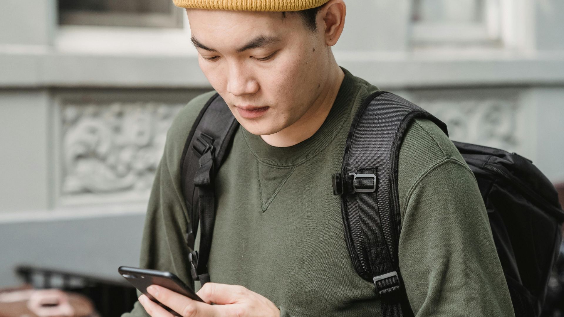 An Asian man wearing casual clothing checks his phone outdoors. City street setting.