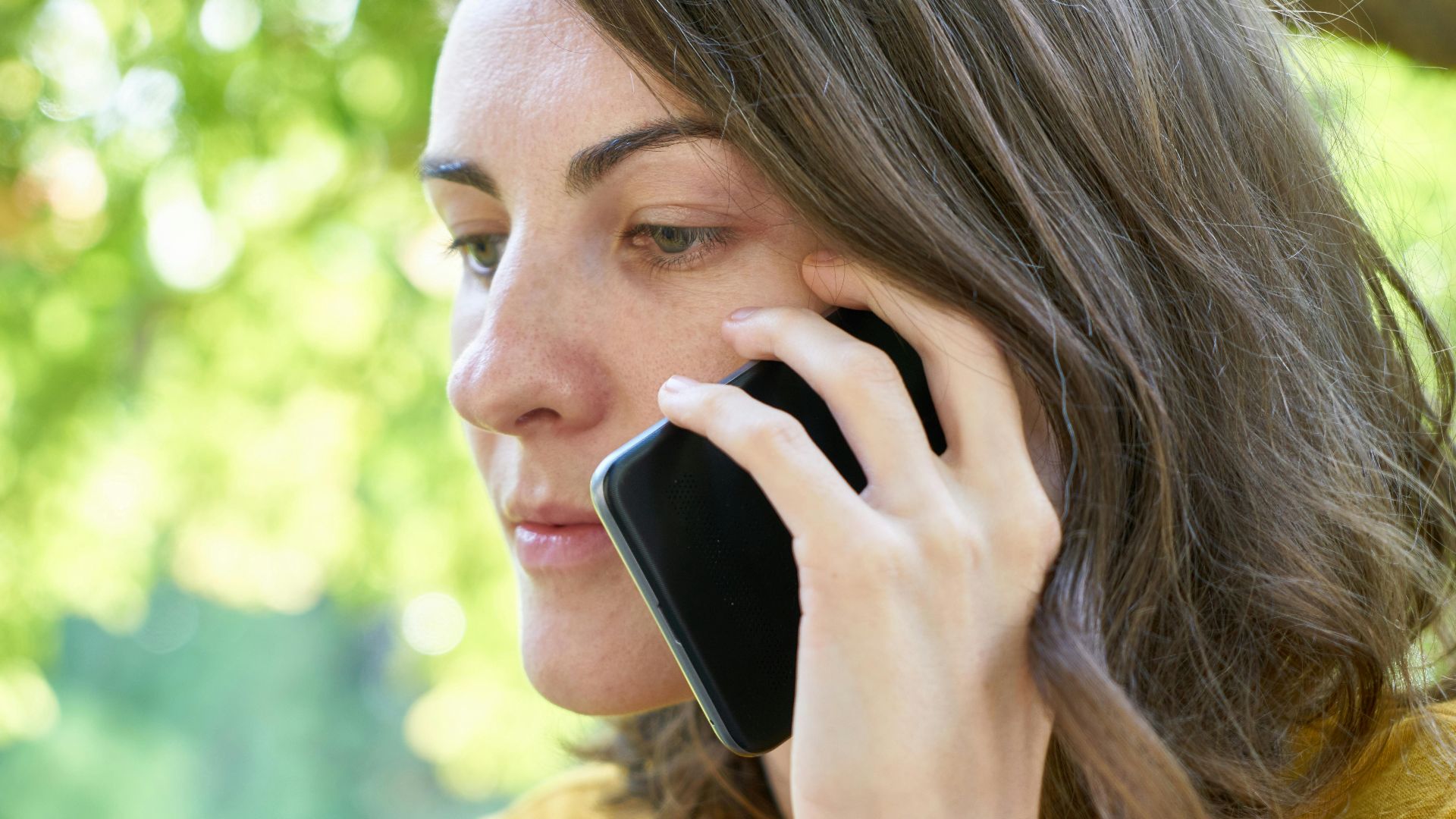 Side profile of a woman using a smartphone outdoors in a sunlit park, wearing a yellow top.