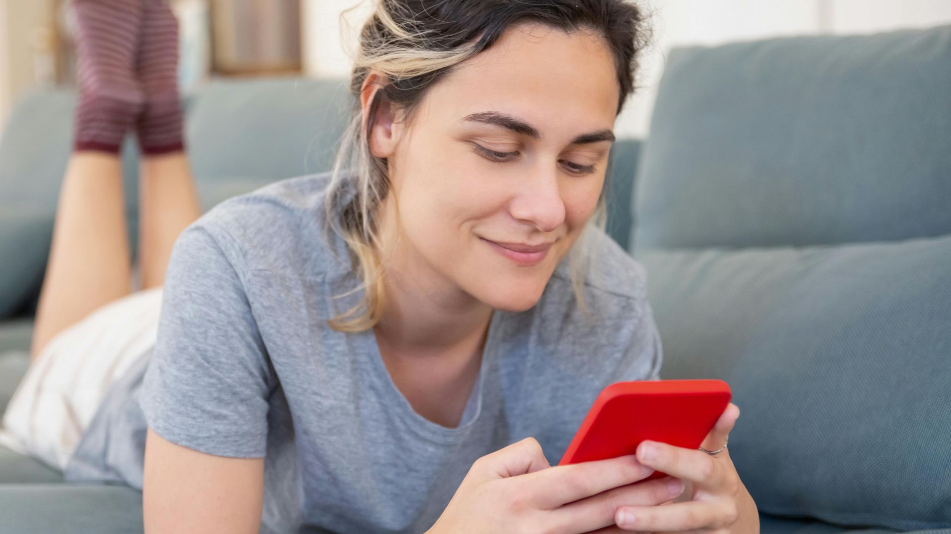 Smiling young woman using a smartphone while lying on a sofa at home, enjoying leisure time.