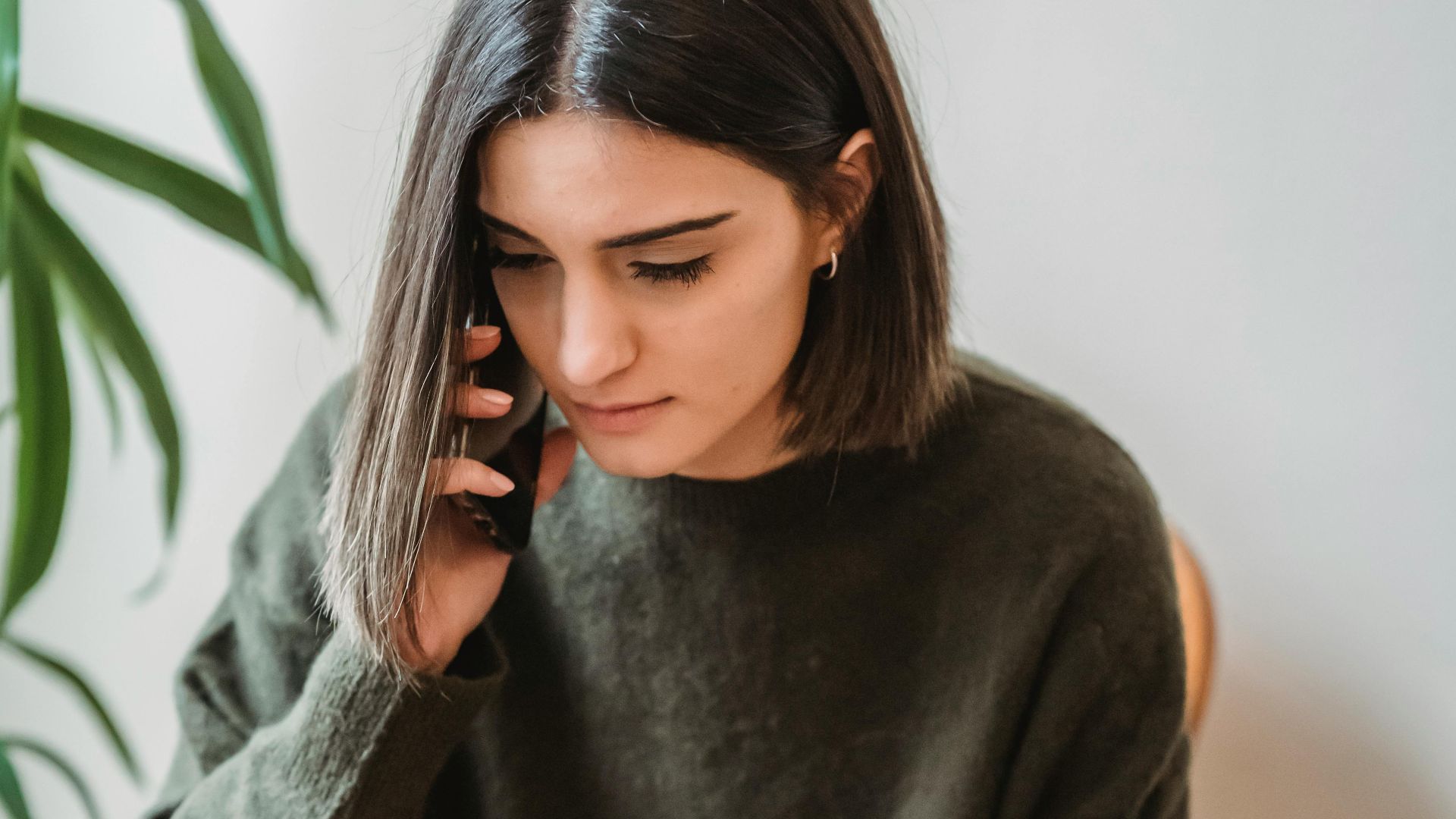 Full body of young ethnic lady with dark hair in casual outfit sitting on chair with crossed legs and talking on smartphone at home