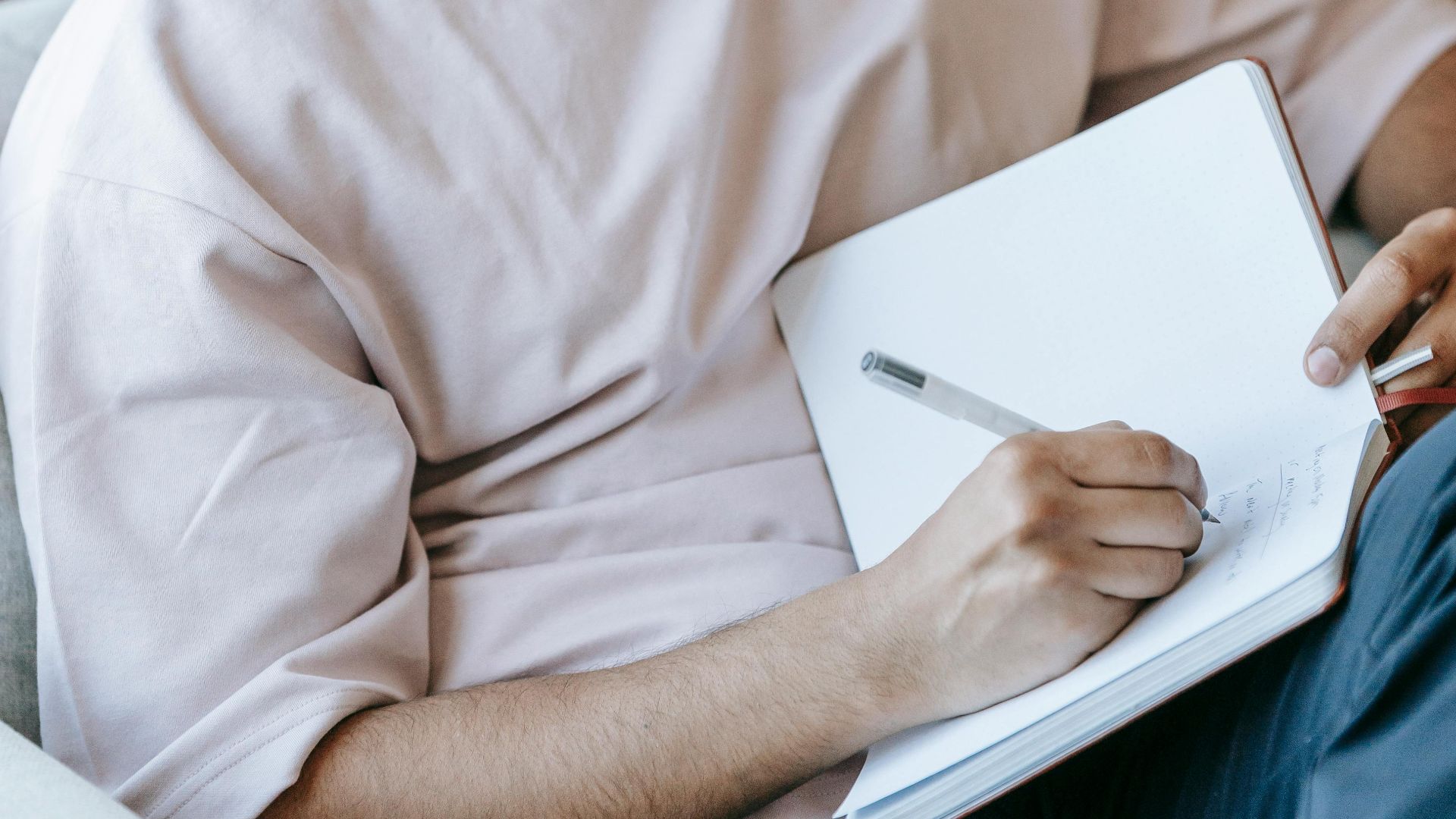 Pensive ethnic male taking notes in diary and listening to music via earbuds while sitting with legs crossed in soft armchair