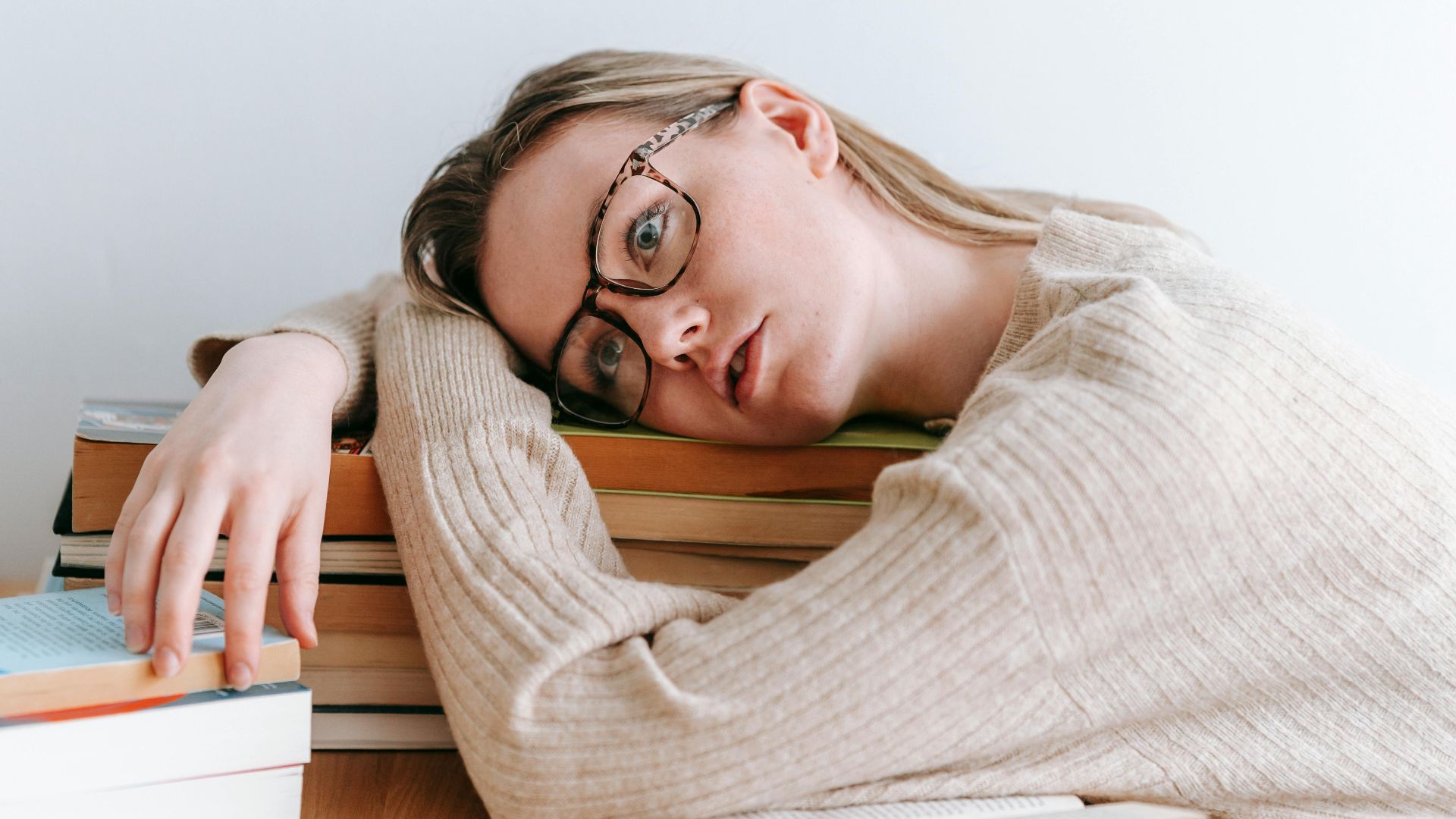 Tired female student in casual clothes lying on table with stack of books and looking away in light room in daytime