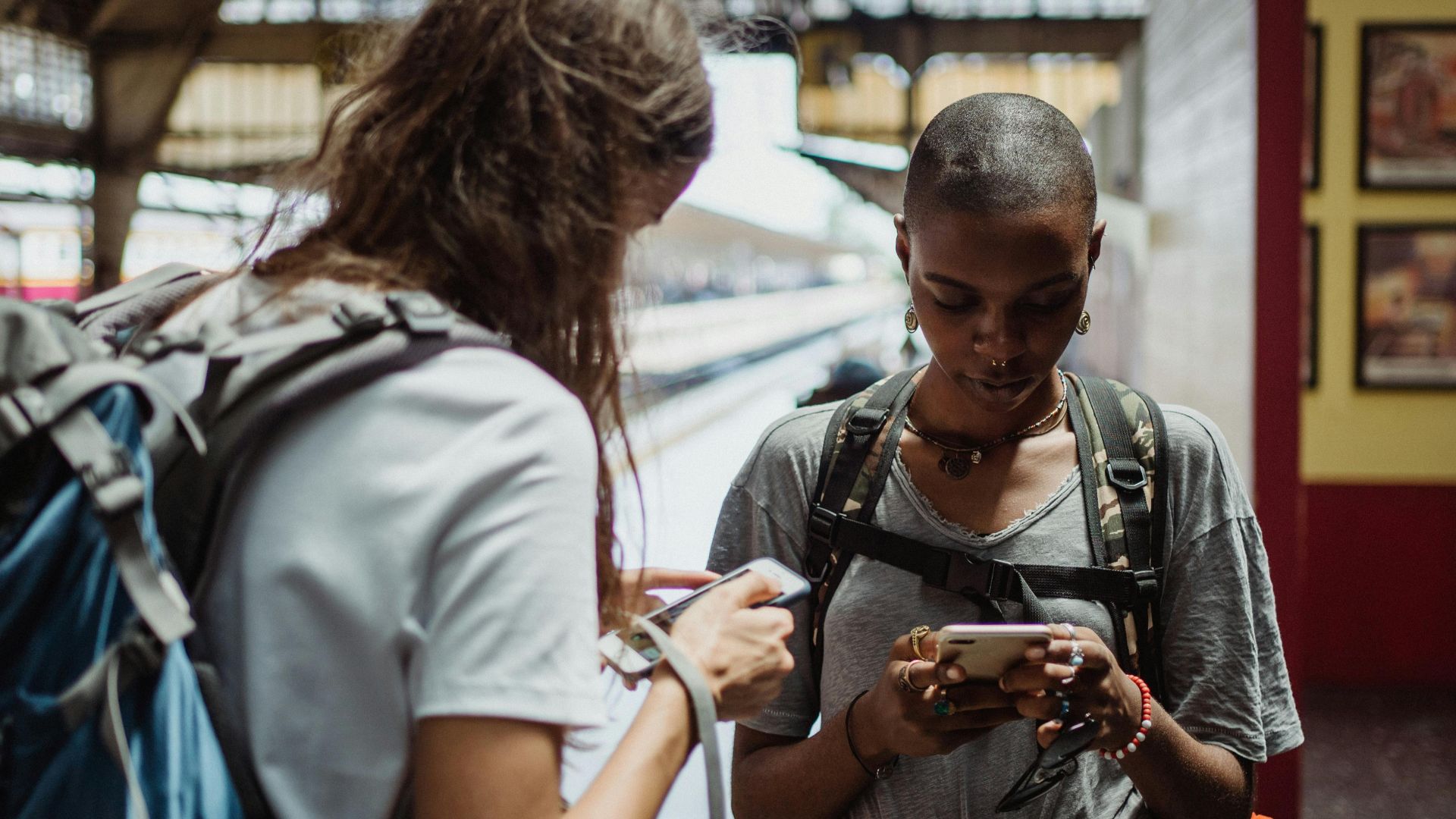 Two travelers using smartphones while waiting at a railway station, backpacks on their shoulders.