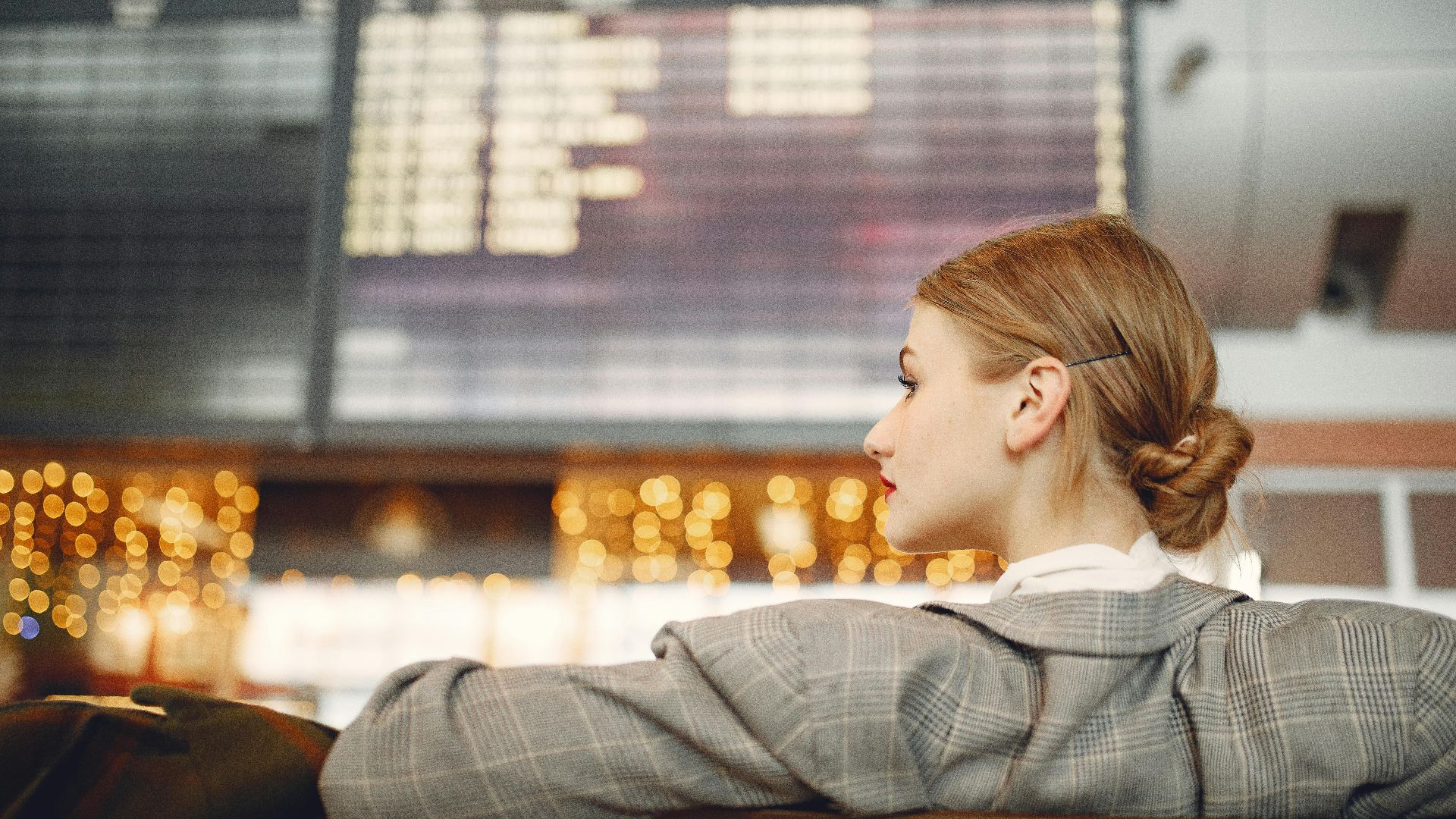 Pensive female designer in checkered jacket waiting for flight near departure board looking away in modern airport in evening