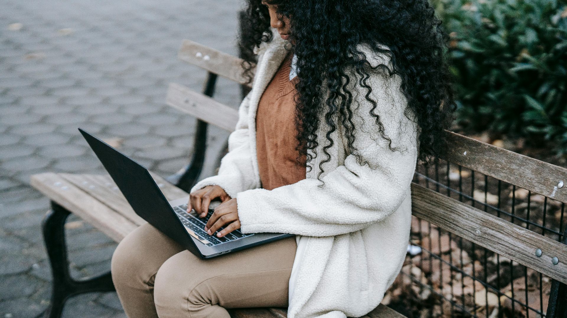 Crop concentrated African American female in warm jacket working on modern netbook and sitting on wooden bench in spring city park
