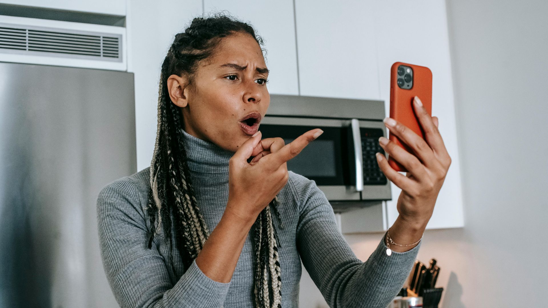 Angry young African American female with long braids in casual clothes pointing at screen nervously while disputing during video call on mobile phone in kitchen