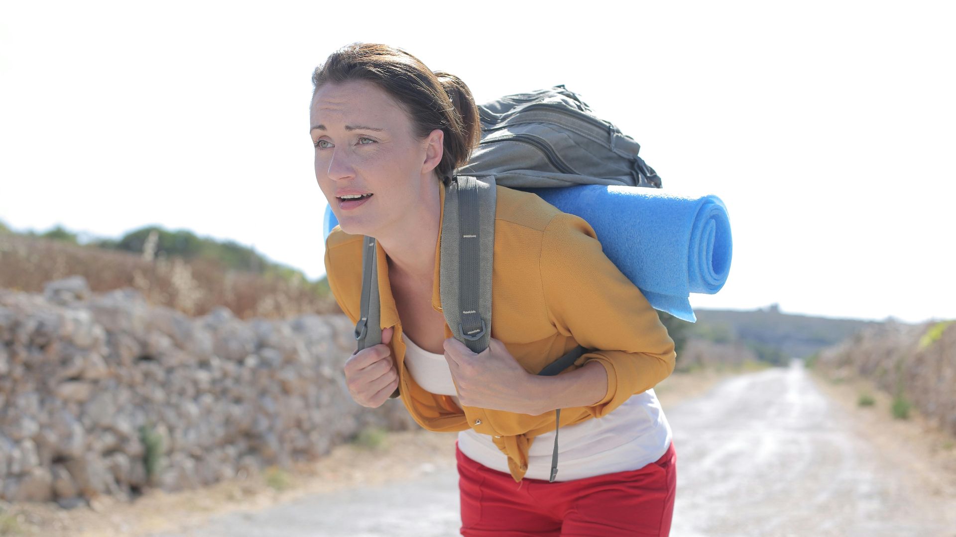 A young woman hiker with a backpack and yoga mat on a sunny road surrounded by nature.