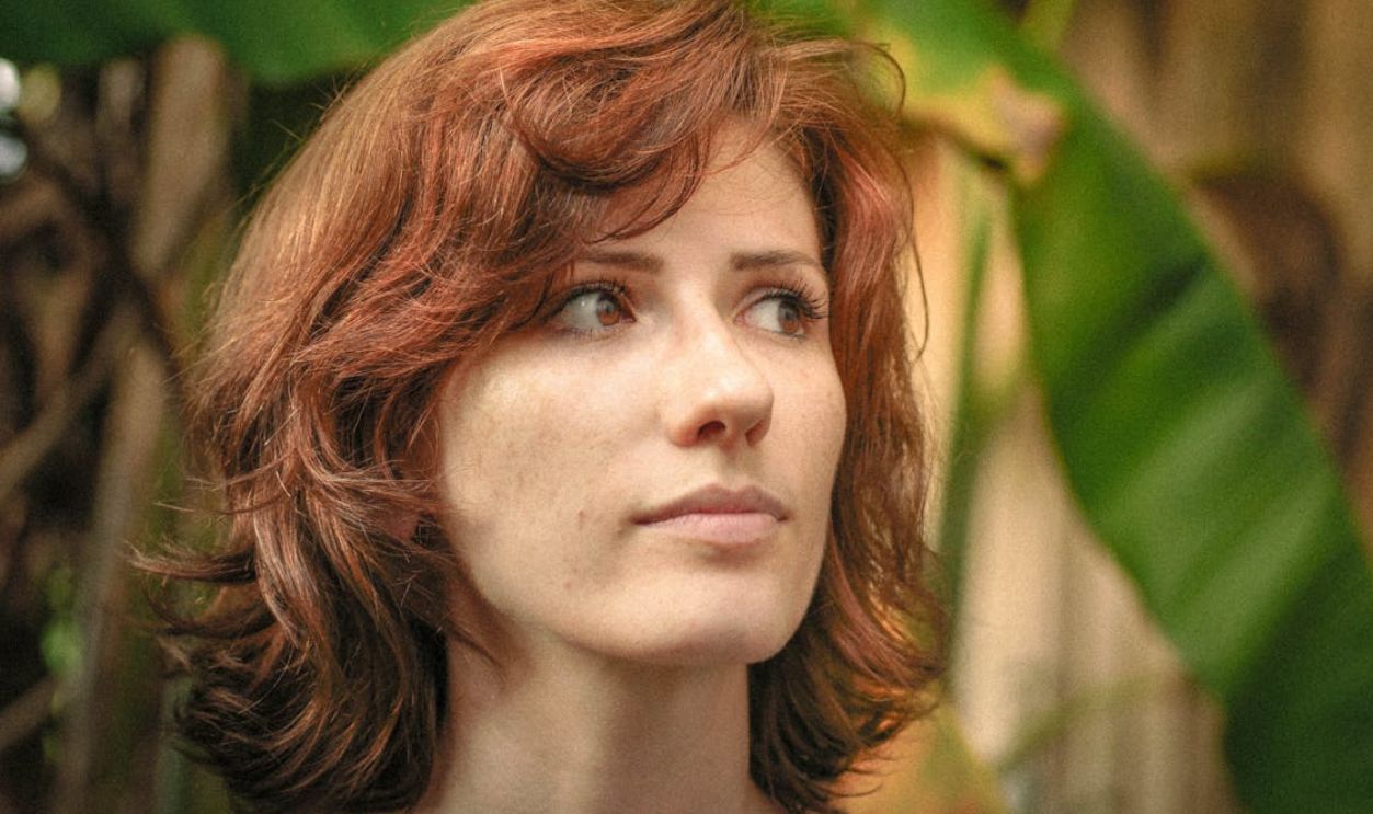 Close-up portrait of a woman with curly hair surrounded by lush green foliage outdoors