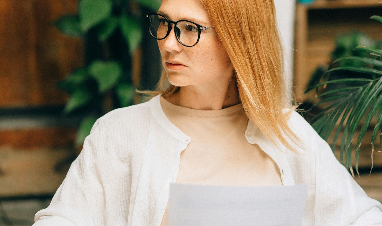 Focused woman with glasses holding documents in a modern workspace, looking thoughtful