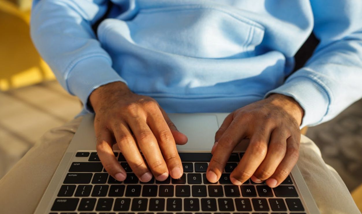 Hands in a blue hoodie typing on a laptop keyboard, indoors, in bright light