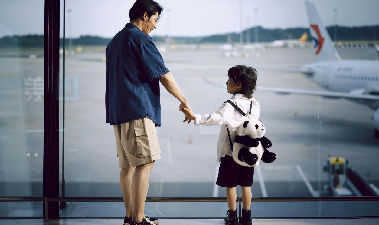 A father and child holding hands, gazing at planes through an airport window
