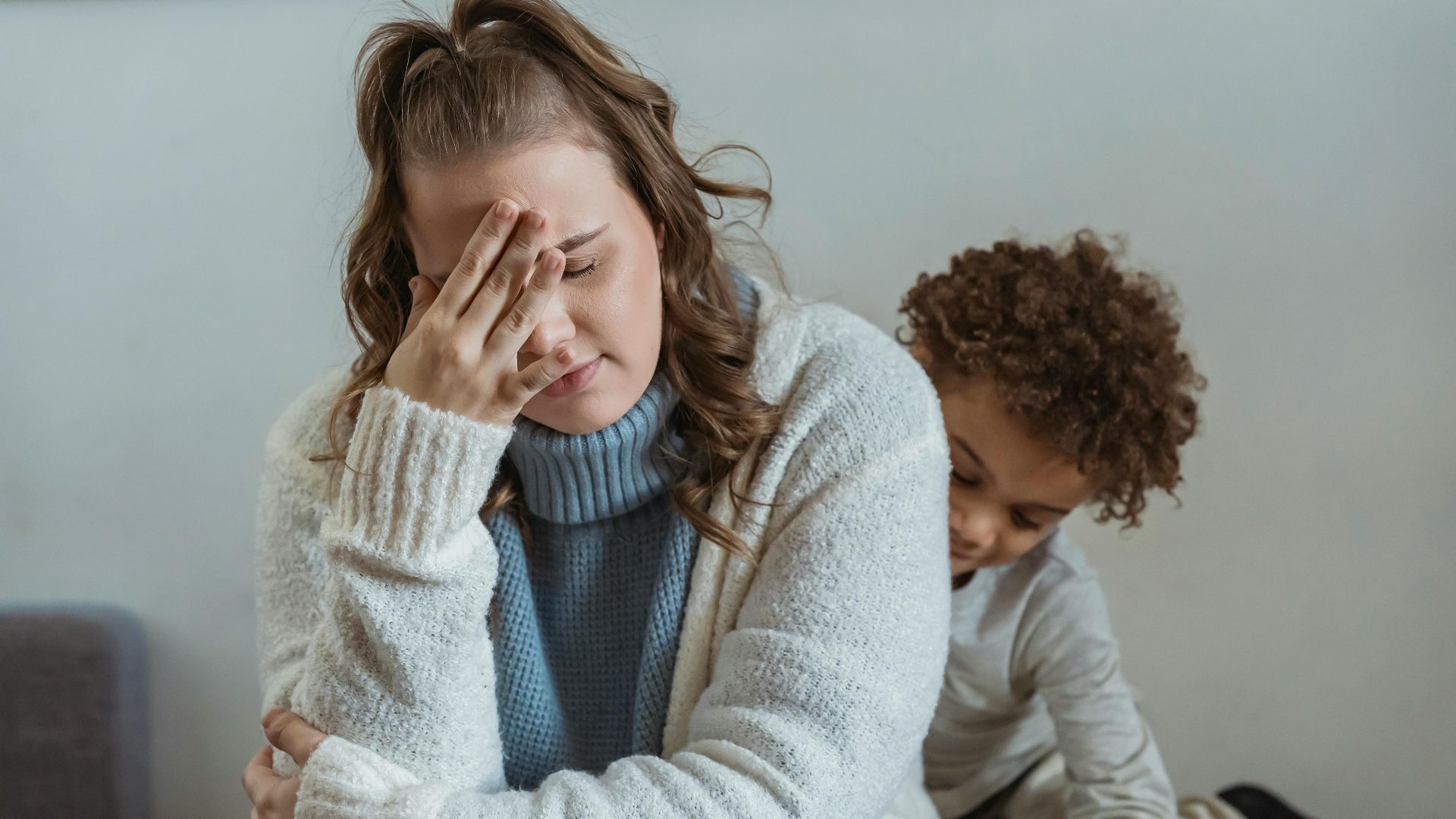 Frustrated mother with hand on forehead and closed eyes sitting near African American son near wall in room at home