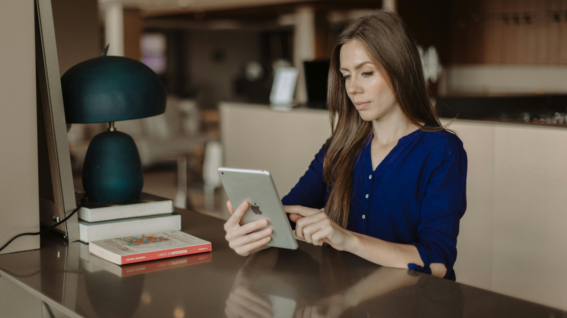 Woman reading on a tablet at a desk with books in a modern office setting.