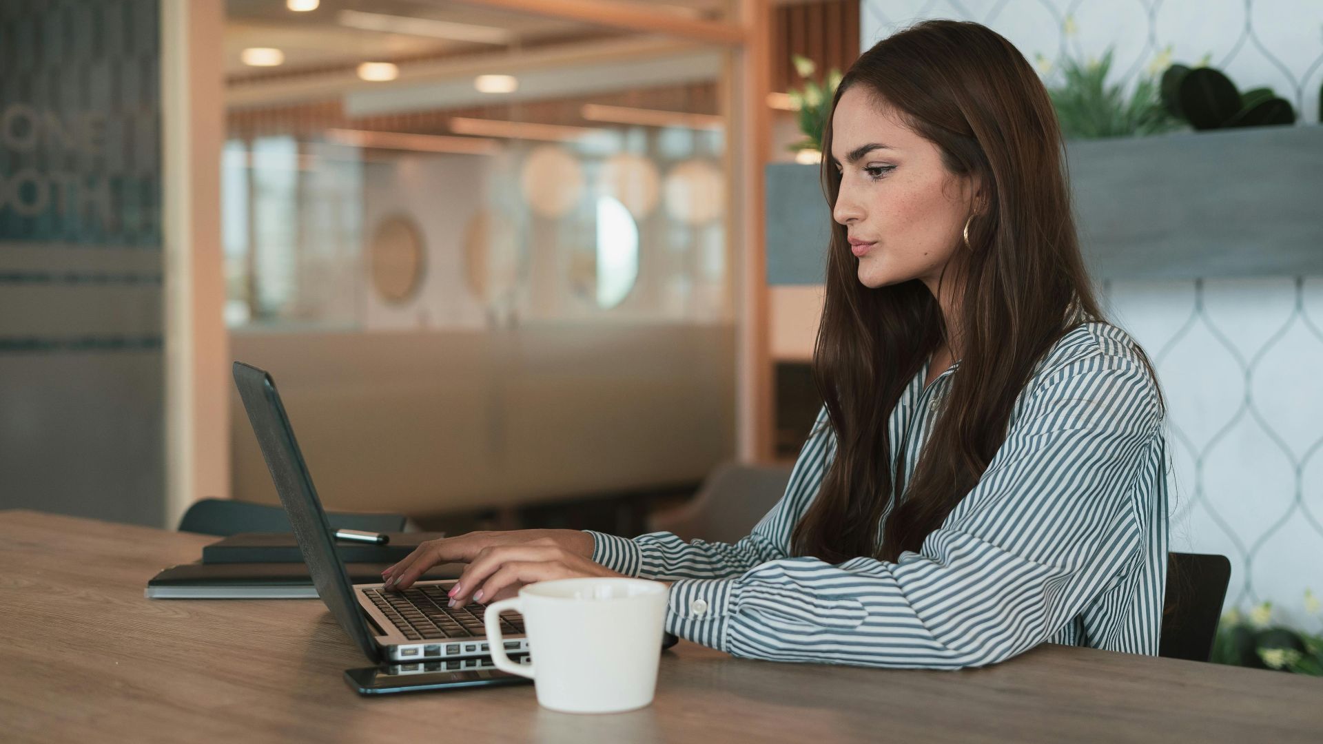 A professional woman working on her laptop at a modern office with a coffee cup nearby.