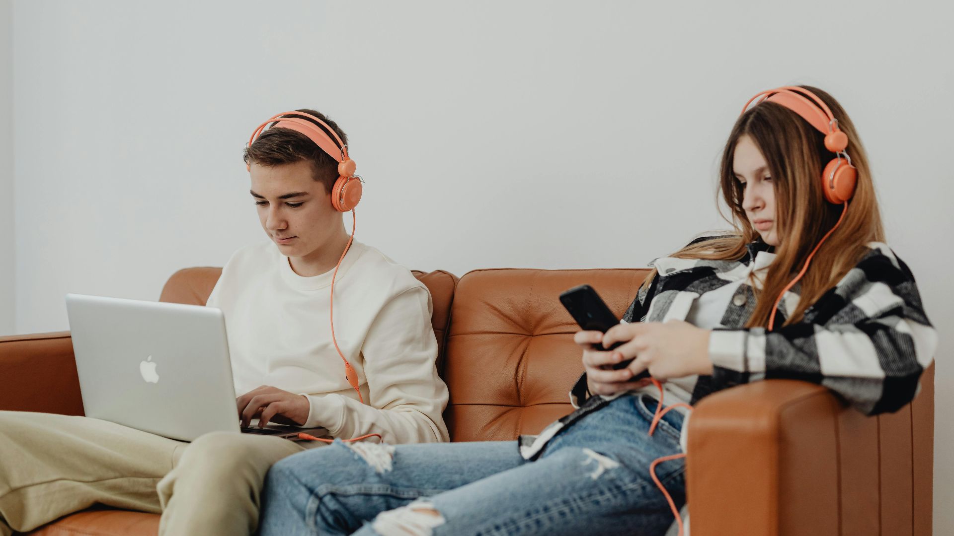 Teens using headphones with laptop and smartphone, lounging on a sofa.