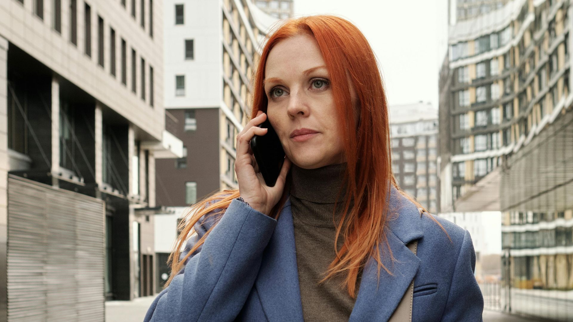 Stylish woman wearing a blue coat talking on a smartphone while walking in a modern cityscape.