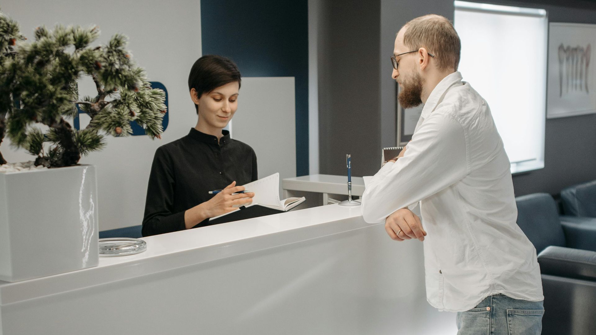 A man and woman at a sleek office reception desk, discussing paperwork in a professional setting.