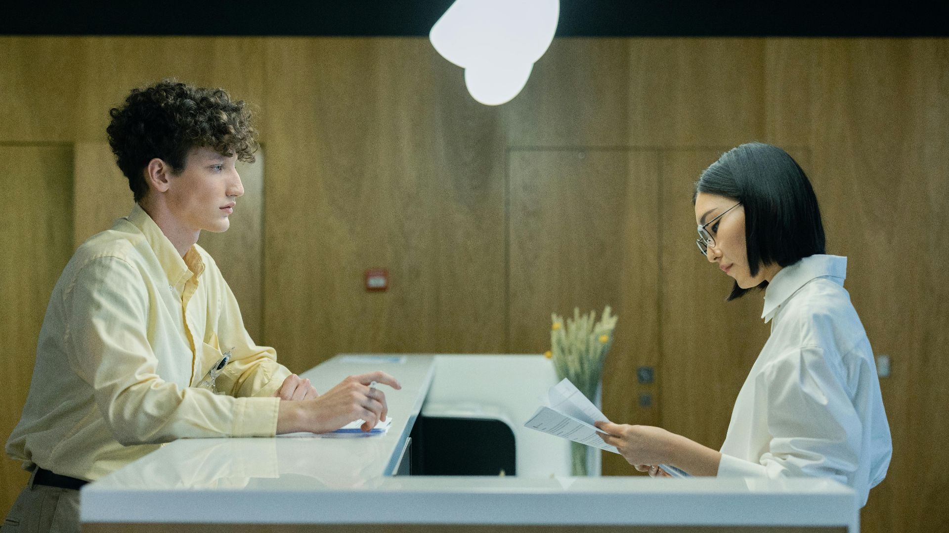 A young man and woman interacting at a modern office reception desk indoors.