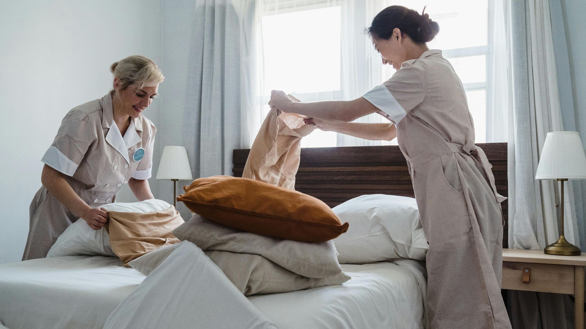 Two housekeepers in uniform fixing a hotel room bed with fresh linen and pillows.