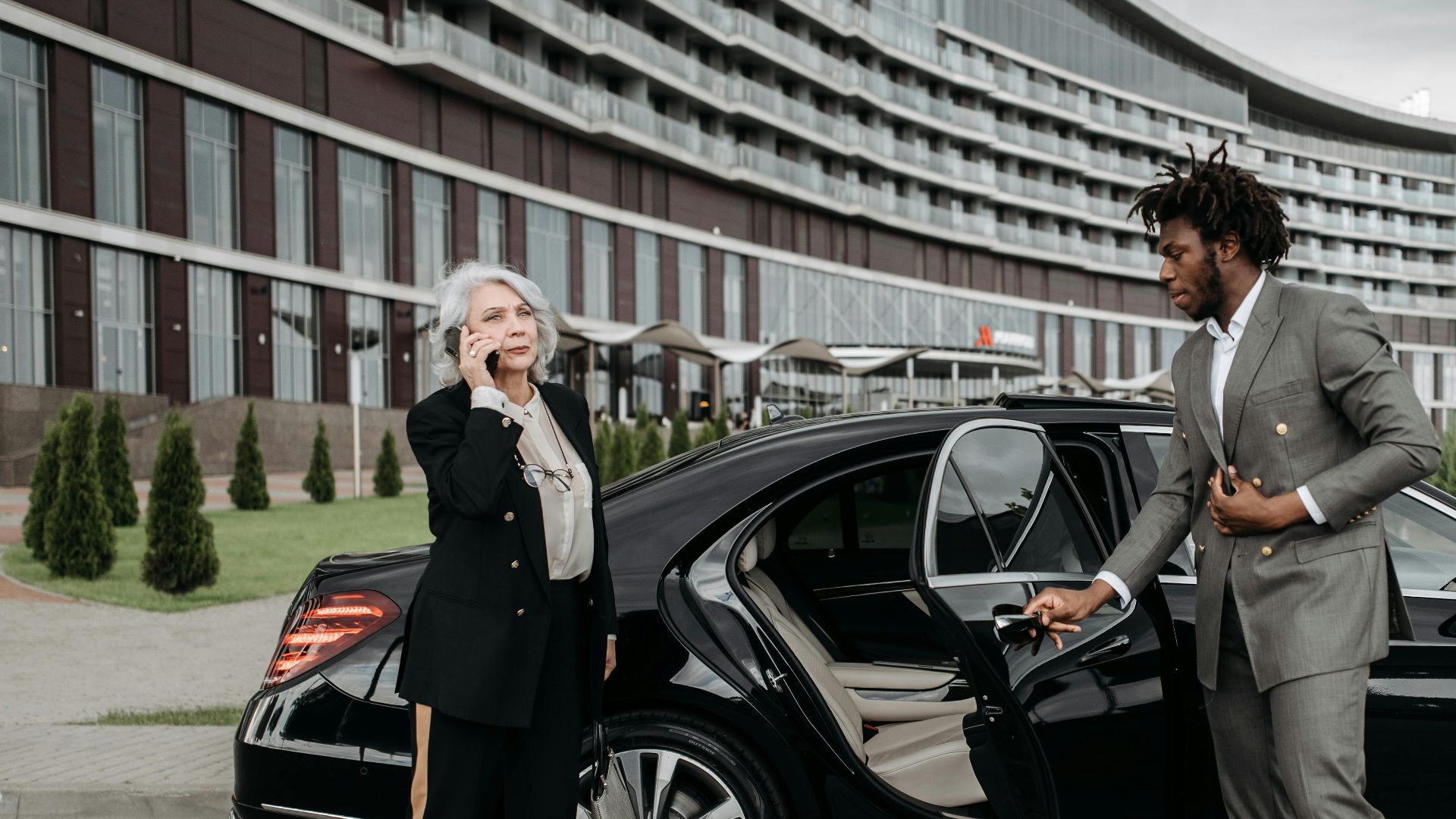 Senior female executive on phone with chauffeur holding car door open, modern architecture backdrop.