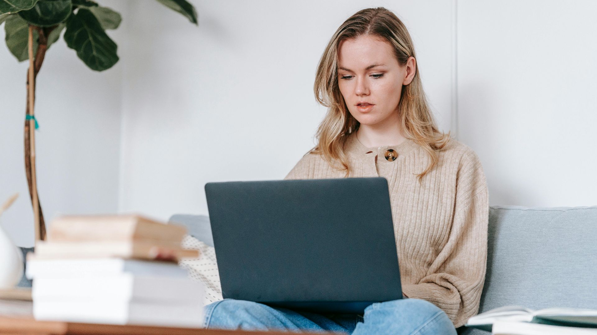 Concentrated young woman browsing internet on portable computer while sitting on couch against books in house on white background