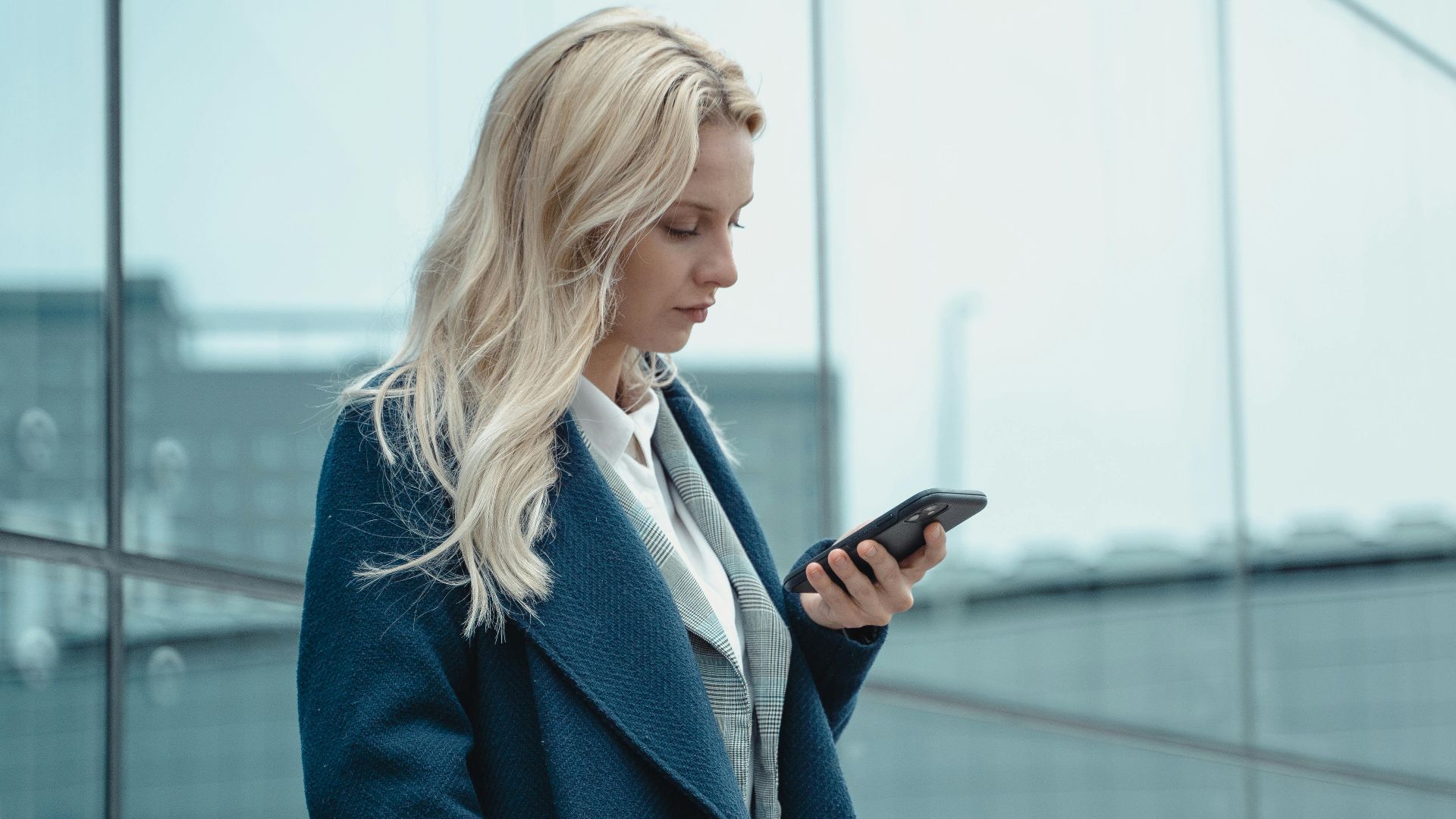 Professional woman in blue coat using smartphone outside office building.