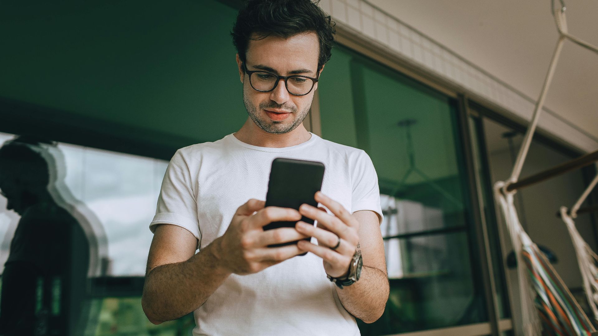 Young man standing on a balcony, using a smartphone, reflecting in the window.