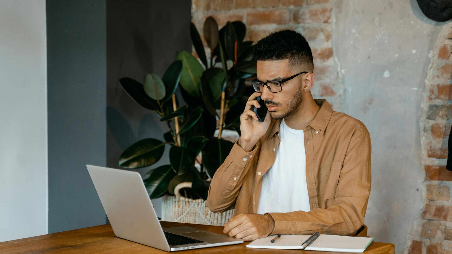 Young man in glasses working on a laptop and phone in a modern office setting.