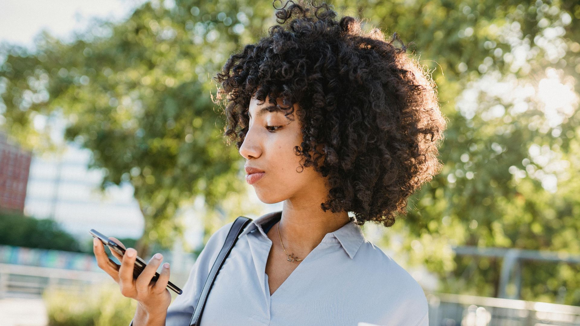 A woman with curly hair holds a coffee cup and checks her smartphone outdoors during daytime.