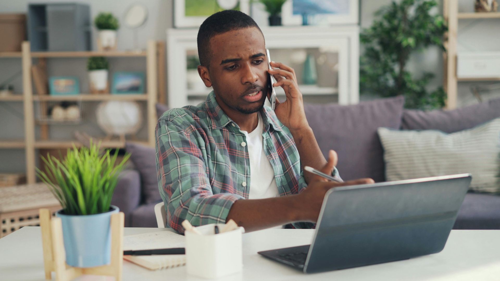 Young man multitasking with phone and laptop in a cozy home office environment.