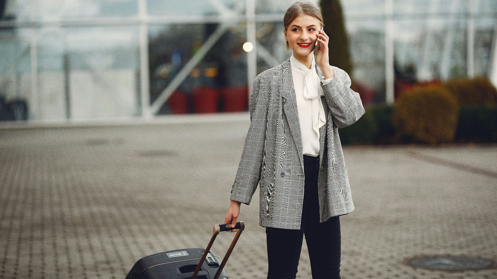 Positive female in trendy outfit talking on phone and looking away while waiting for taxi with suitcase near airport terminal