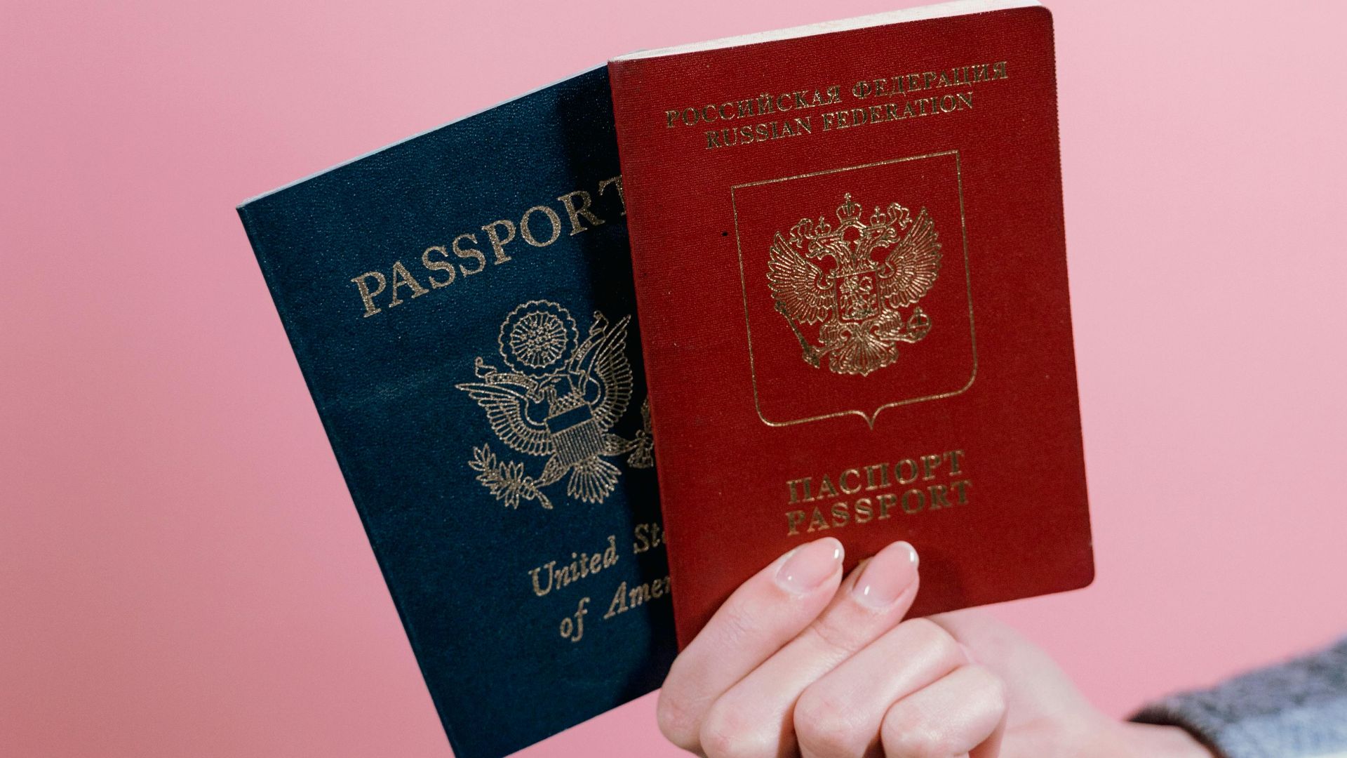 Close-up of two passports held by a hand against a pink background.