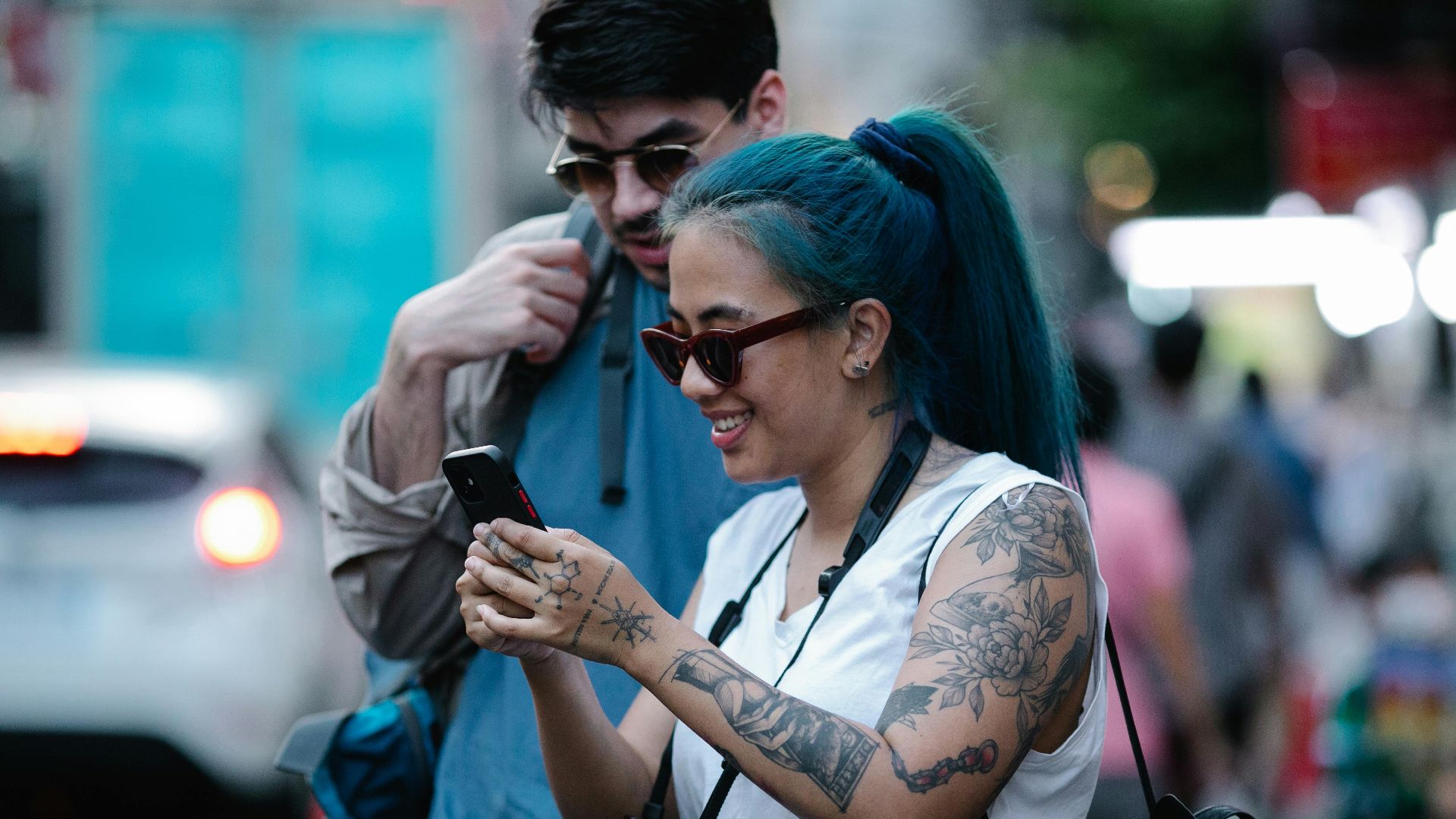 Two young adults with tattoos explore urban streets, capturing moments on a mobile phone.