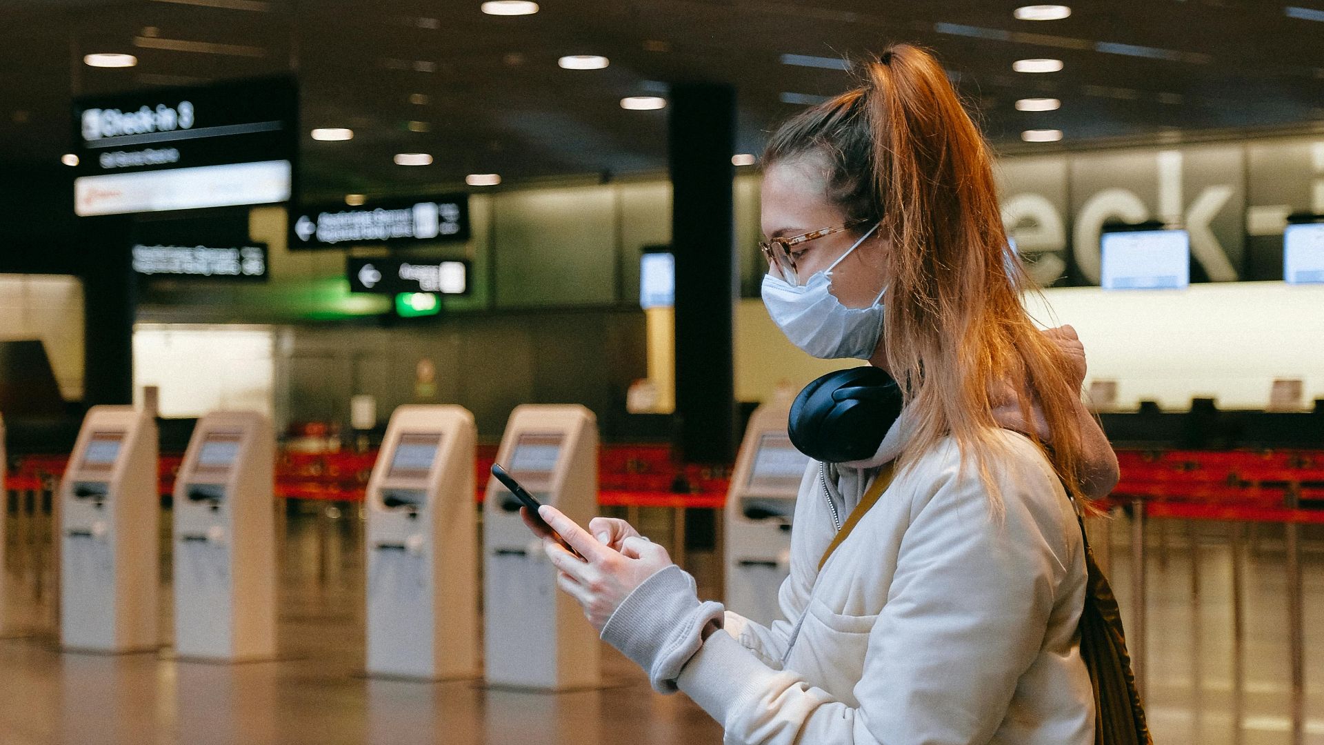 Woman wearing a mask, texting in airport terminal with signage in background.