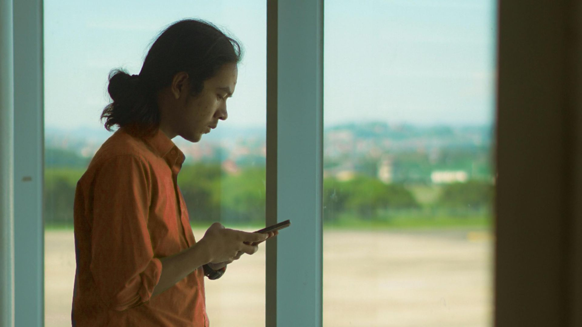 Man in orange shirt using a smartphone while standing at an airport window, overlooking the runway.