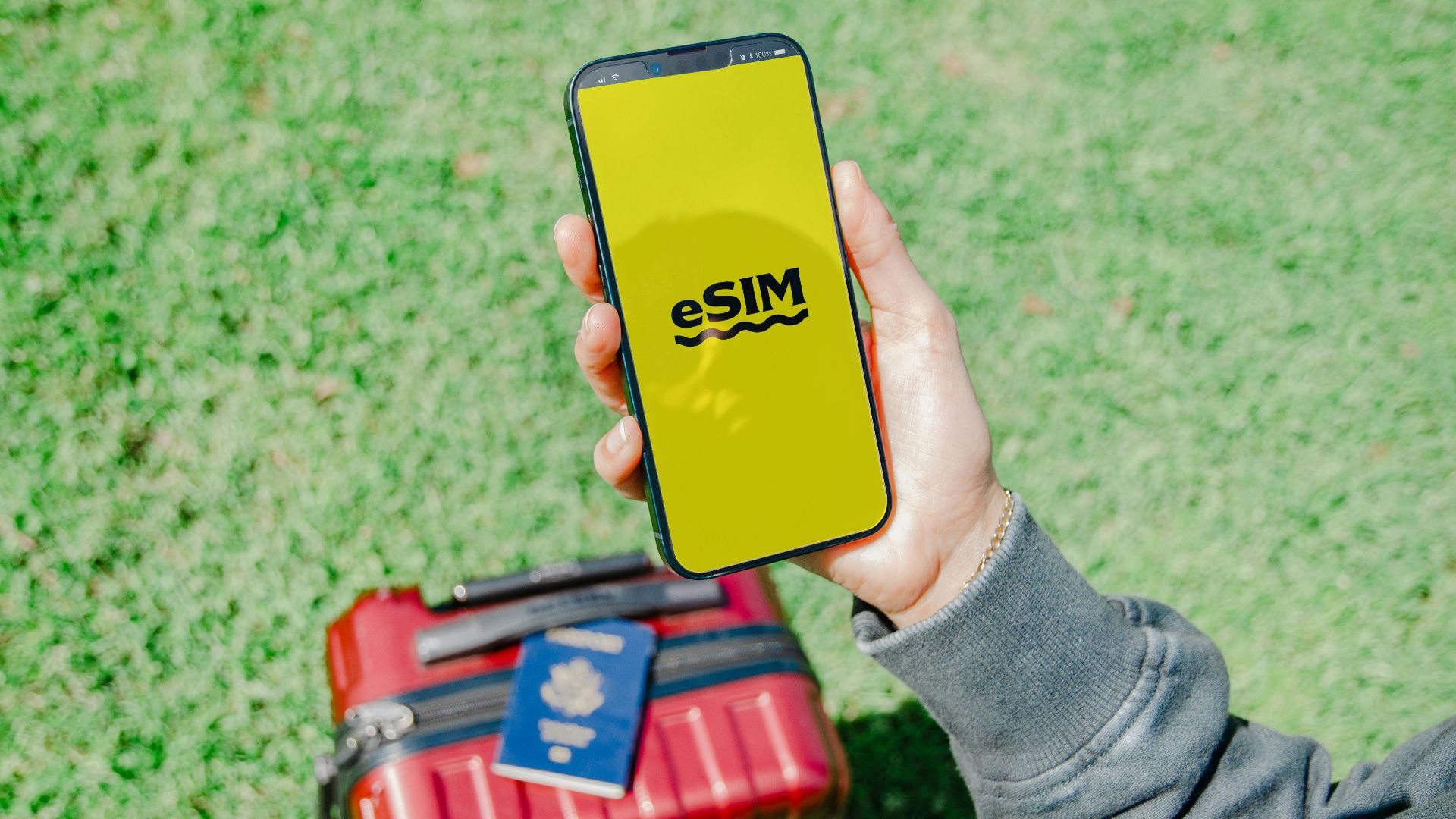 Traveler in Los Angeles holding a smartphone with eSIM near a red suitcase on grass.