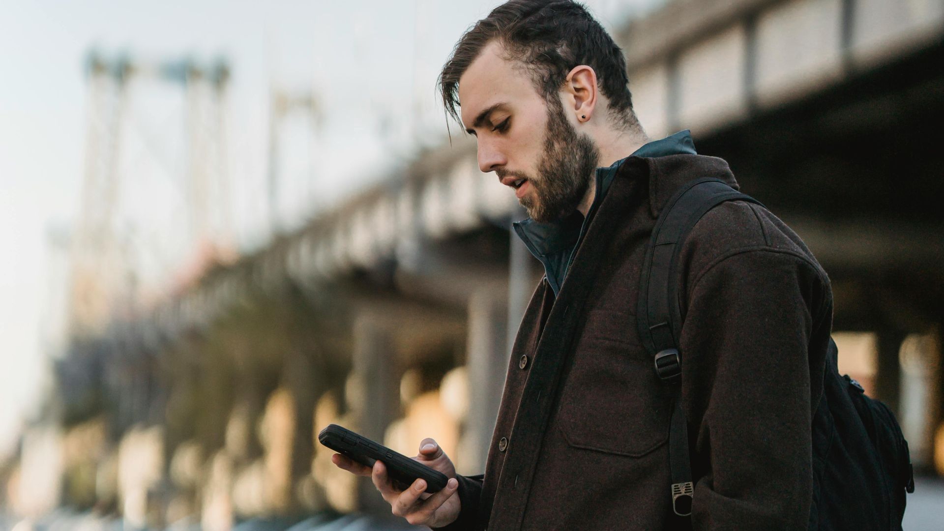 Side view of serious young man in casual clothes standing on asphalt road while text messaging on phone near cars and bridge in city street in daytime