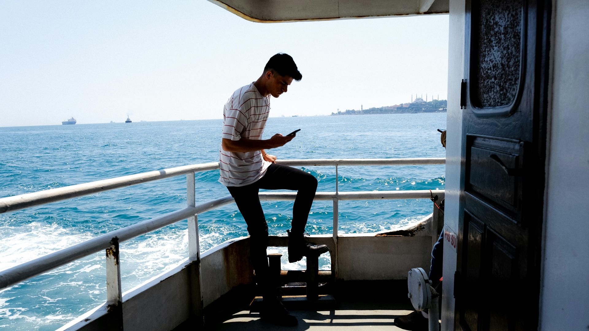A man stands on a boat, looking at his phone with a scenic sea backdrop on a sunny day.