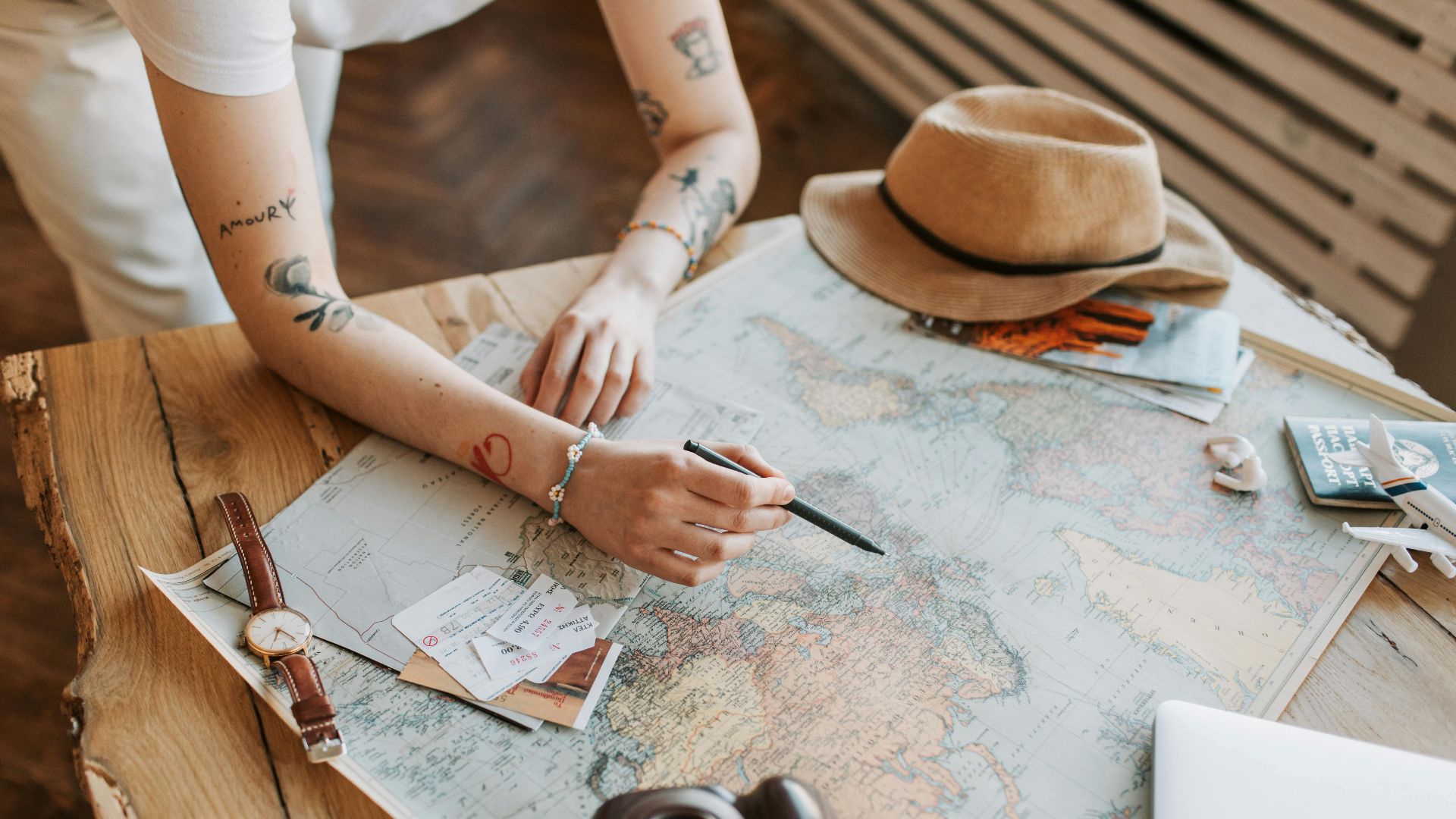 Tattooed woman planning a trip with a world map, passports, and travel essentials on a wooden table.