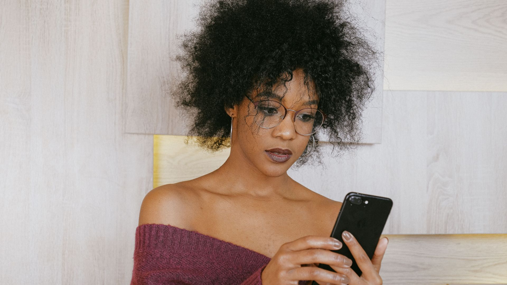 A fashionable woman with curly hair using her smartphone indoors, wearing glasses and a cozy sweater.