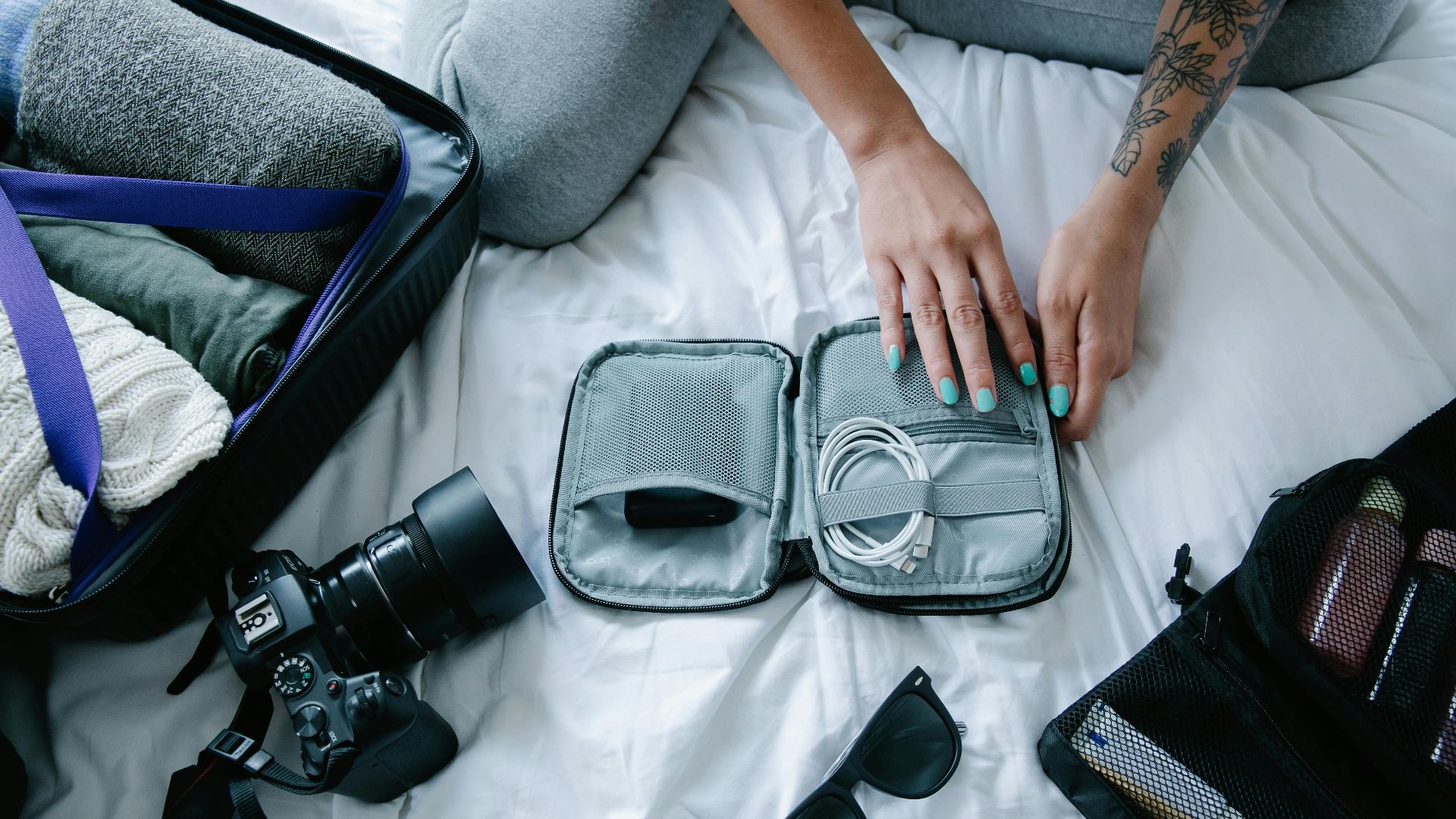 Woman with tattoos packs electronics, camera, and sunglasses in a suitcase on a bed.