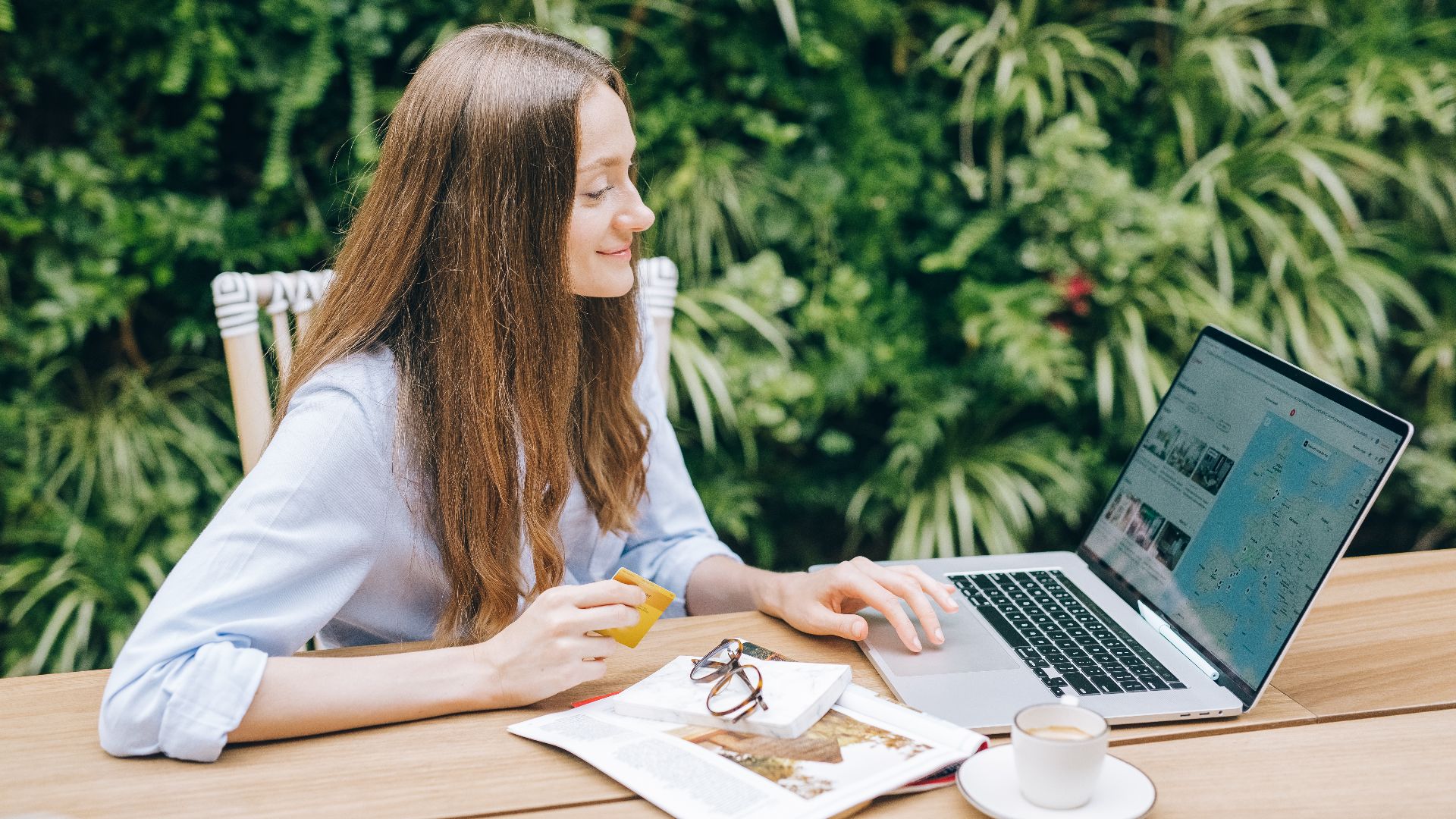 Young woman sitting at a table with a laptop and coffee, enjoying outdoor workspace.