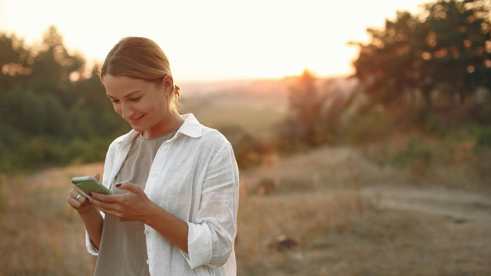 Cheerful female in casual clothes smiling while enjoying surfing smartphone in rural landscape in summer sunny evening