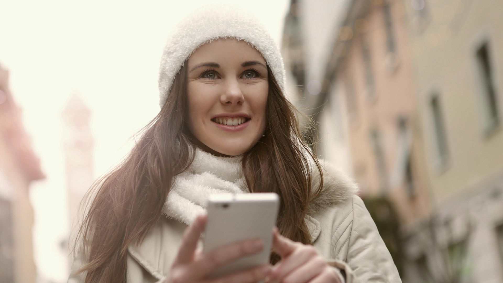 A cheerful woman in winter clothing uses a smartphone outdoors.