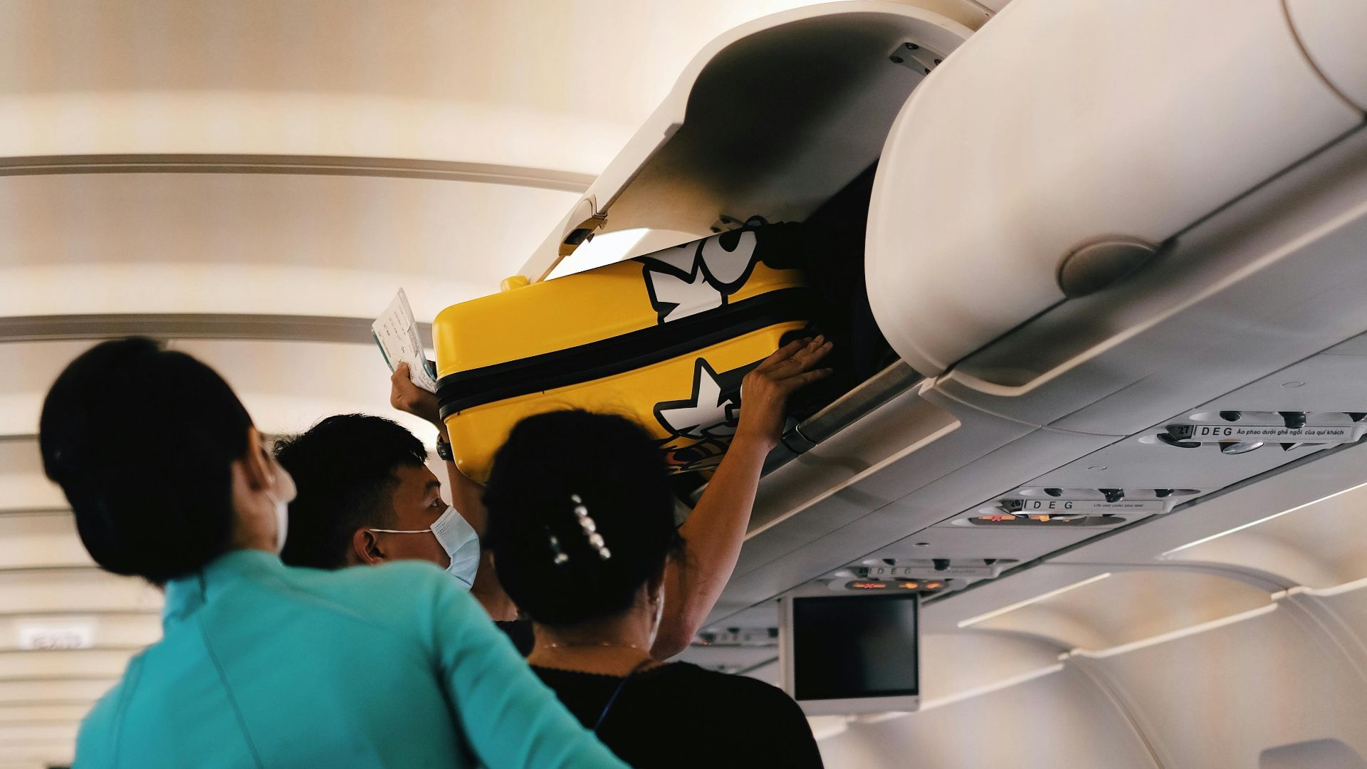 Passengers placing a yellow suitcase in the overhead bin of an airplane cabin. Interior with crew assistance.