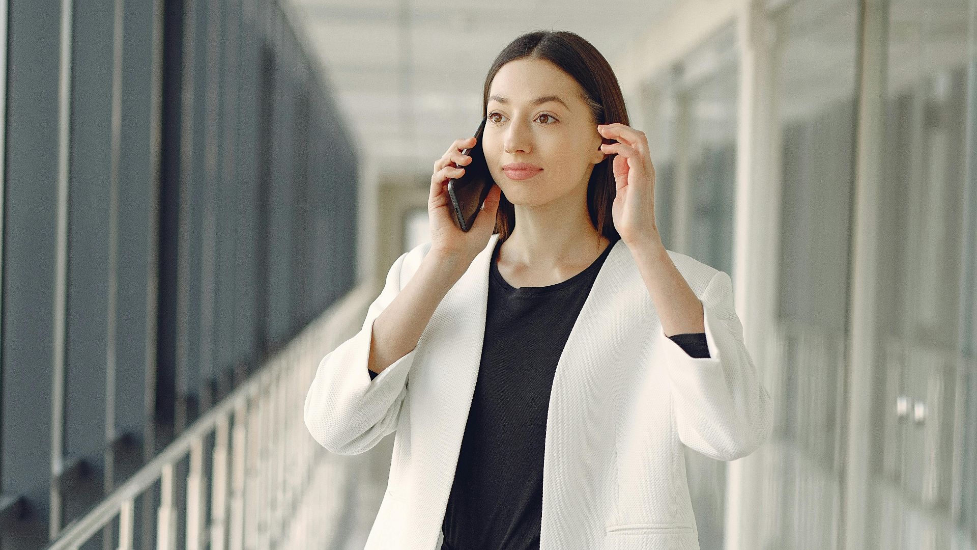 Positive young businesswoman in stylish formal wear with suitcase talking on smartphone and standing in modern airport corridor while waiting for flight